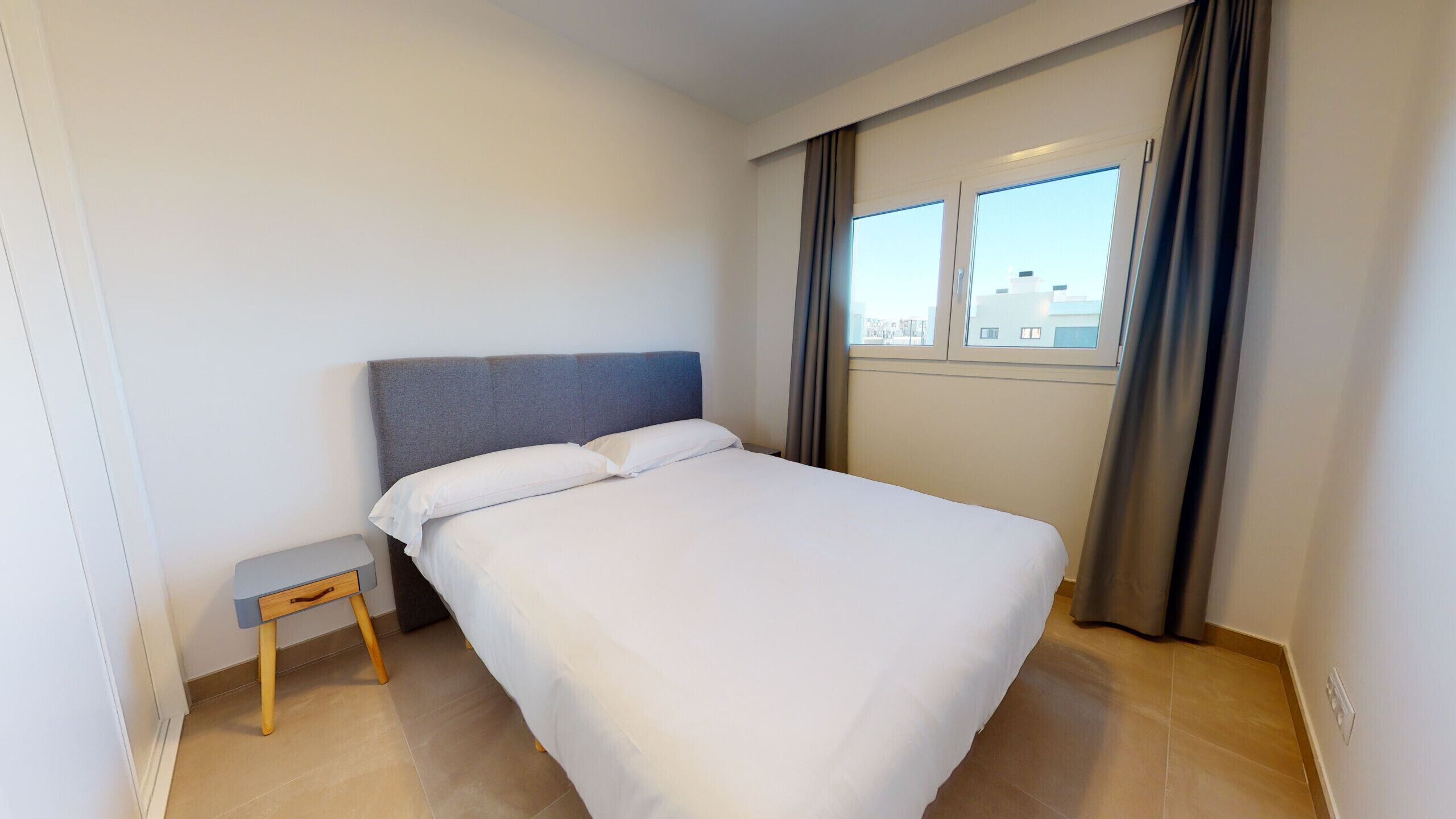 A neatly made bed in a hotel room with a small wooden nightstand, a window with curtains, and a view of buildings outside.