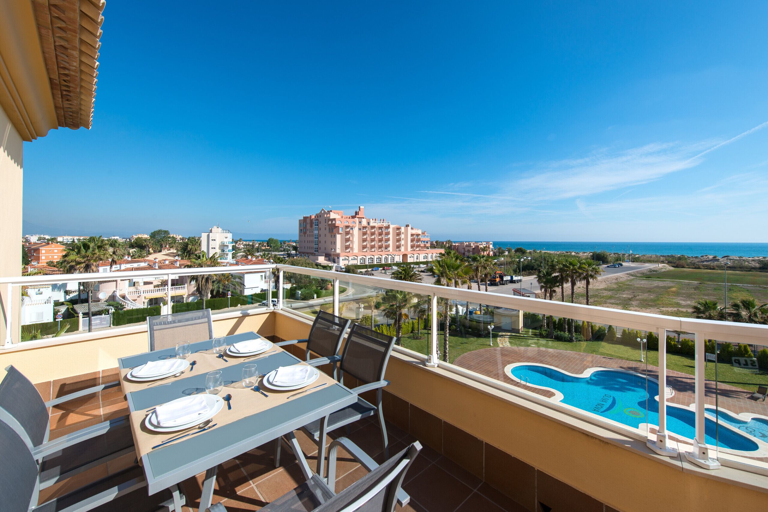 A balcony with a dining table set for four, overlooking a swimming pool and a view of a coastal town.
