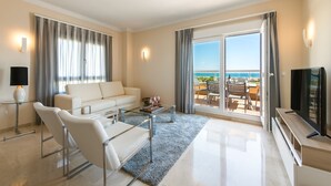 Una sala de estar moderna con sofá, sillones, una mesa de centro y televisión. Cuenta con ventanas amplias con cortinas, un balcón con mobiliario exterior y vistas al mar.