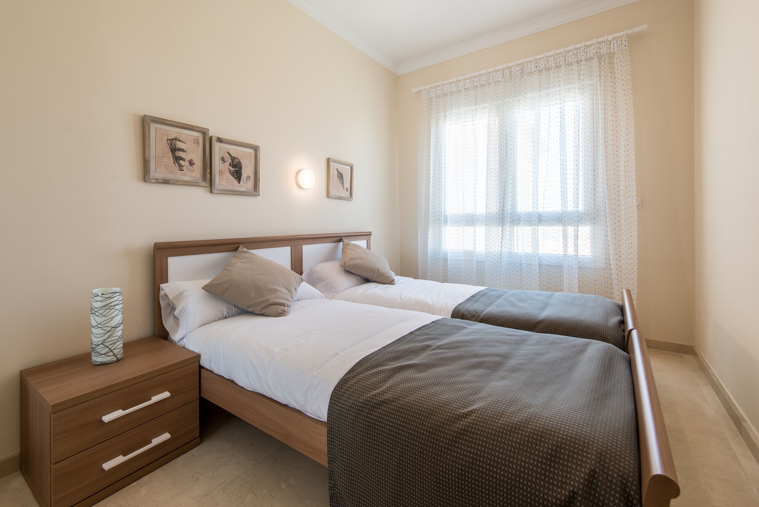 A double bed with a wooden headboard, two bedside tables with drawers, a large window with sheer curtains, and three framed pictures on the wall.
