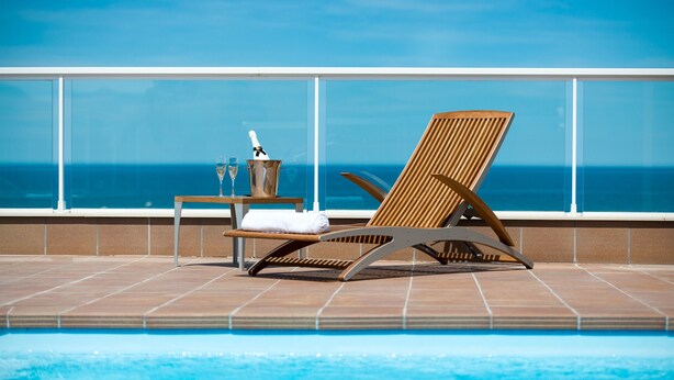 Zona de piscina con una silla de descanso, una mesa con botella y vaso, y vista al océano.