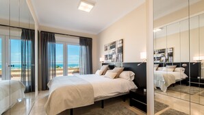 Un dormitorio moderno con una cama grande, una mesita de noche, un espejo y vistas al mar a través de amplios ventanales.