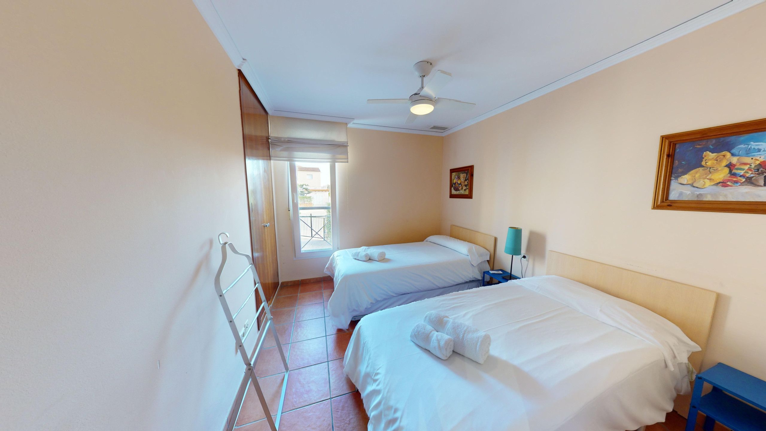 A hotel room with two beds, a ceiling fan, a painting, and a view through a window.