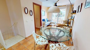 Un coin repas avec une table en verre et des chaises blanches, un canapé à l’arrière-plan et un escalier menant à une autre pièce.