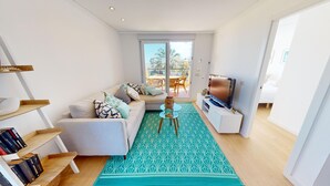 Una sala de estar moderna con un sofá, una mesa de centro y un televisor. Se ve un balcón con mobiliario exterior a través de puertas de vidrio.