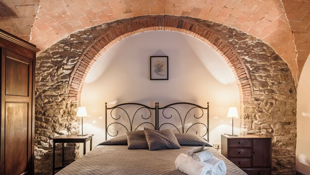 A bedroom with a stone arch ceiling, a bed with a quilted bedspread, two bedside tables with lamps, and a wooden wardrobe. - Studio on organic farm located in ancient hamlet.  (Monterotondo Marittimo)
