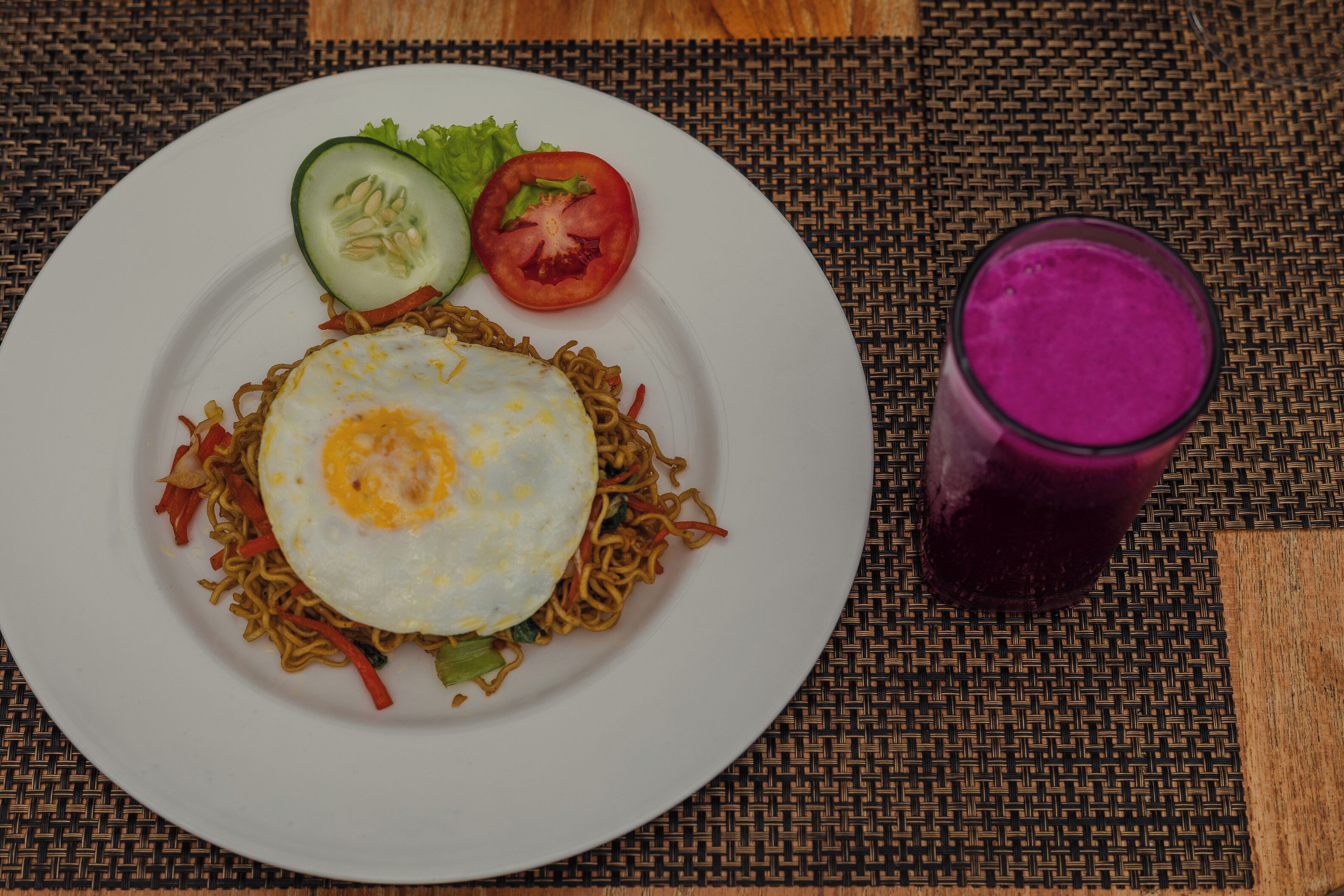Un plato de fideos con un huevo frito, una rodaja de pepino, una rodaja de tomate y un vaso de bebida morada sobre un mantel tejido.