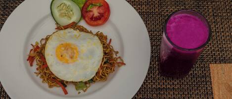 Un plato de fideos con un huevo frito, una rodaja de pepino, una rodaja de tomate y un vaso de bebida morada sobre un mantel tejido.