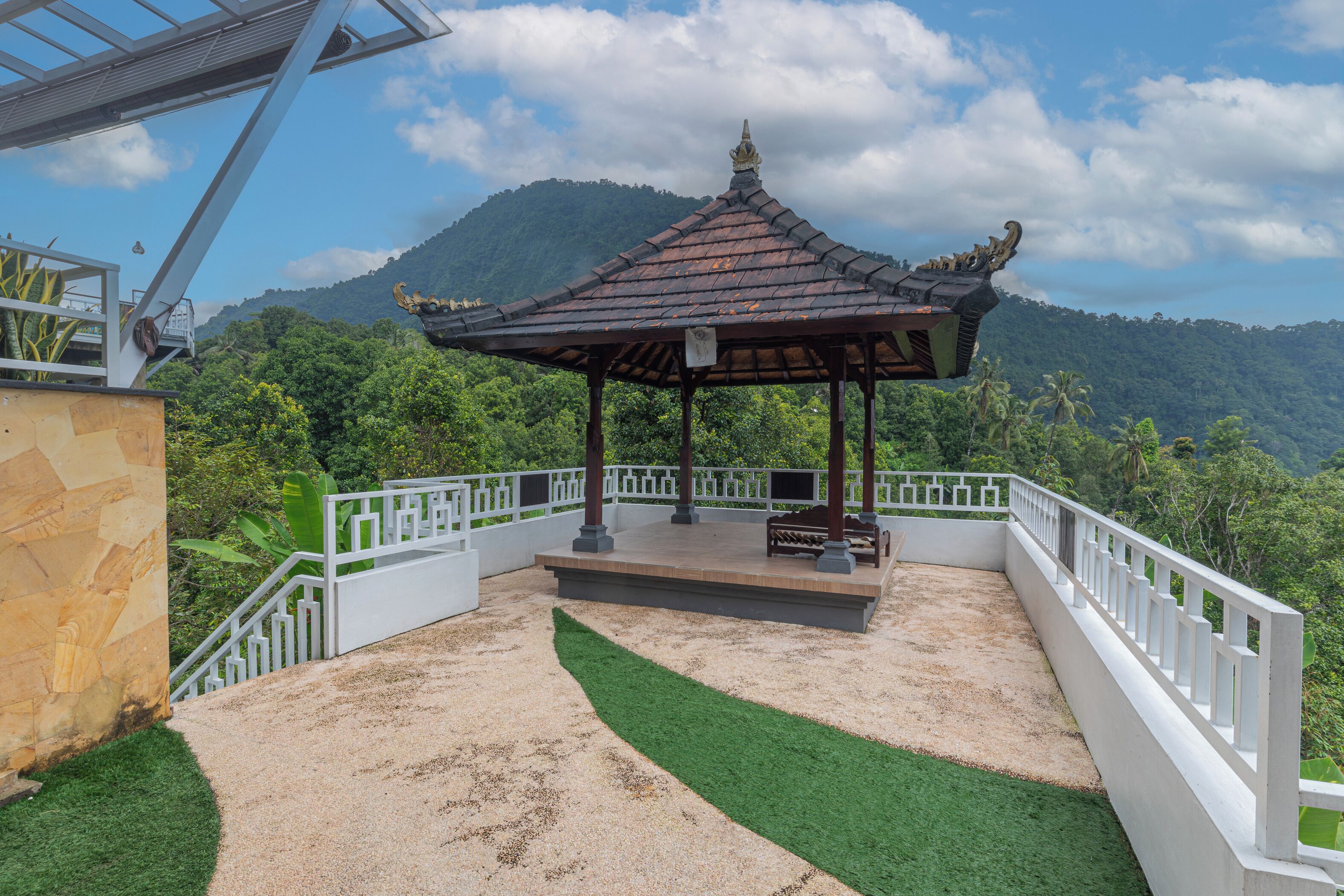 Ein Pavillon mit Ziegeldach und Bank, umgeben von einem weißen Zaun und grünem Rasen, mit Blick auf eine gebirgige Landschaft.