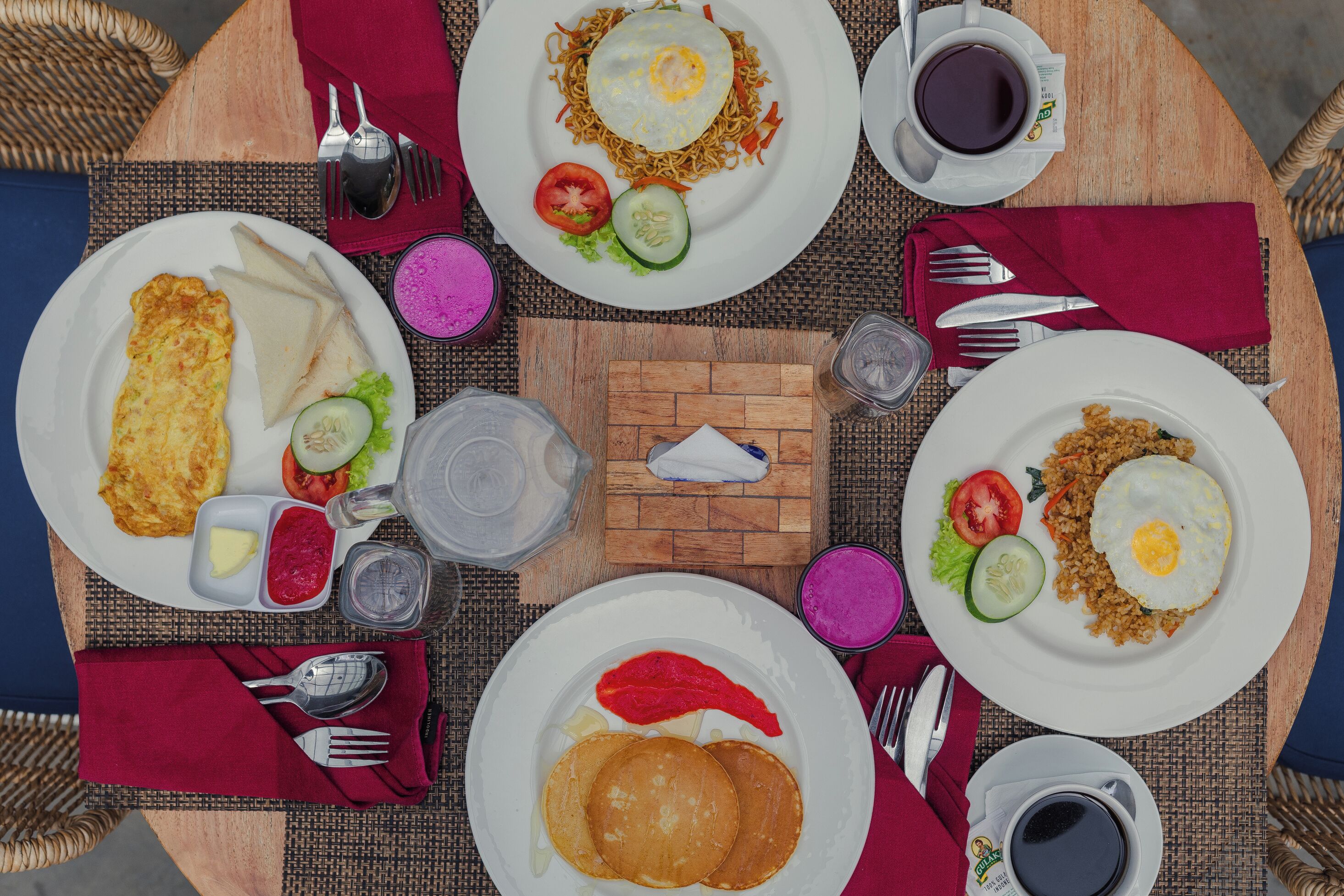 Una mesa dispuesta con diferentes platos de desayuno, incluyendo panqueques, huevos fritos y un plato de fideos, junto con tazas de café y salsas.