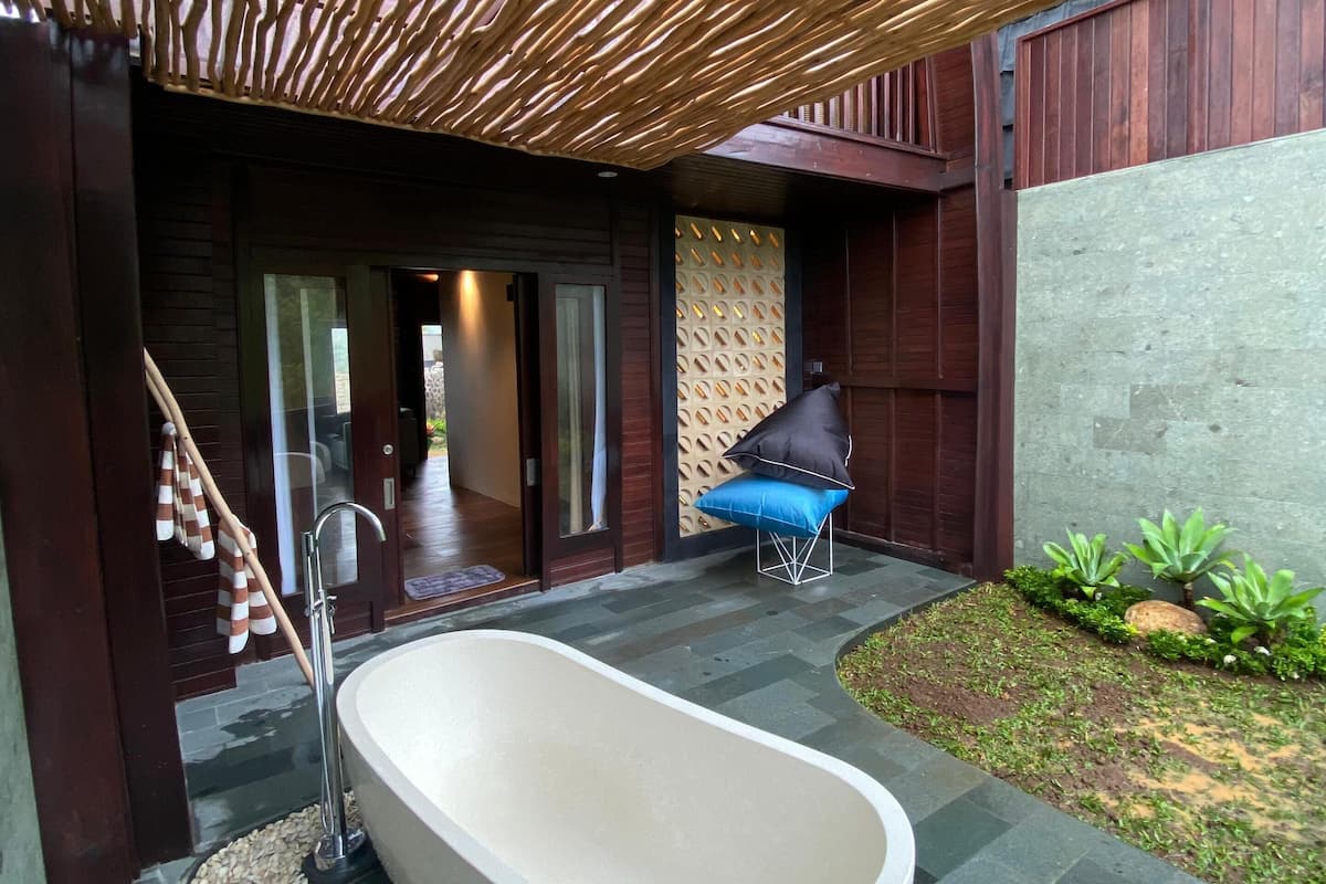 Un baño moderno al aire libre con una bañera grande, una pérgola de madera y una pequeña zona de estar con cojín.