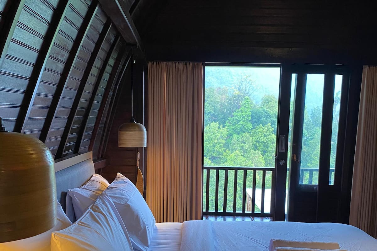 Habitación de hotel con una cama grande, ropa de cama blanca, techo de madera y balcón con vistas a la vegetación.