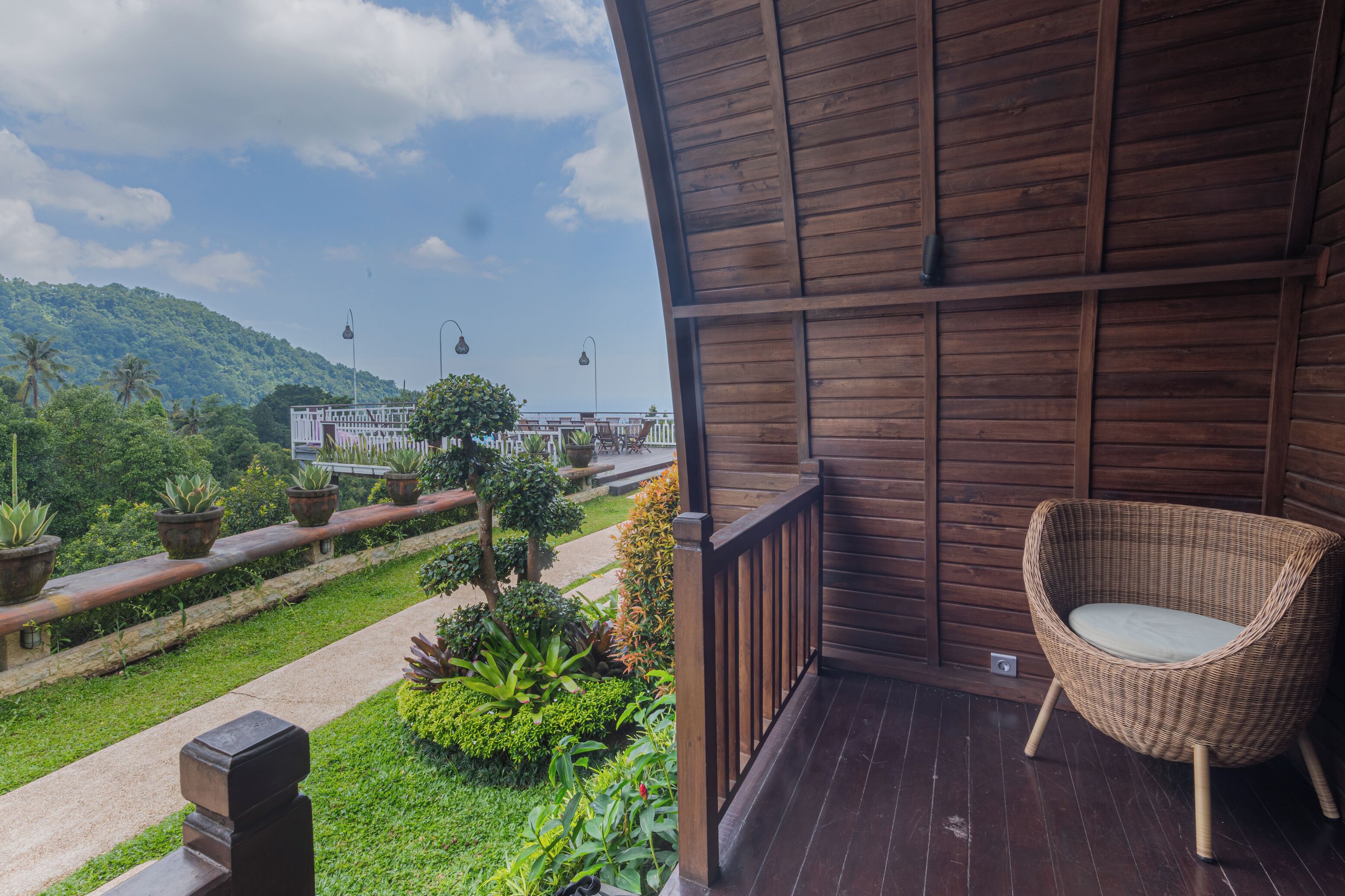 Un balcon en bois agrémenté d’une chaise en osier, de plantes en pot et offrant une vue sur un paysage verdoyant et luxuriant ainsi que sur des bâtiments éloignés.