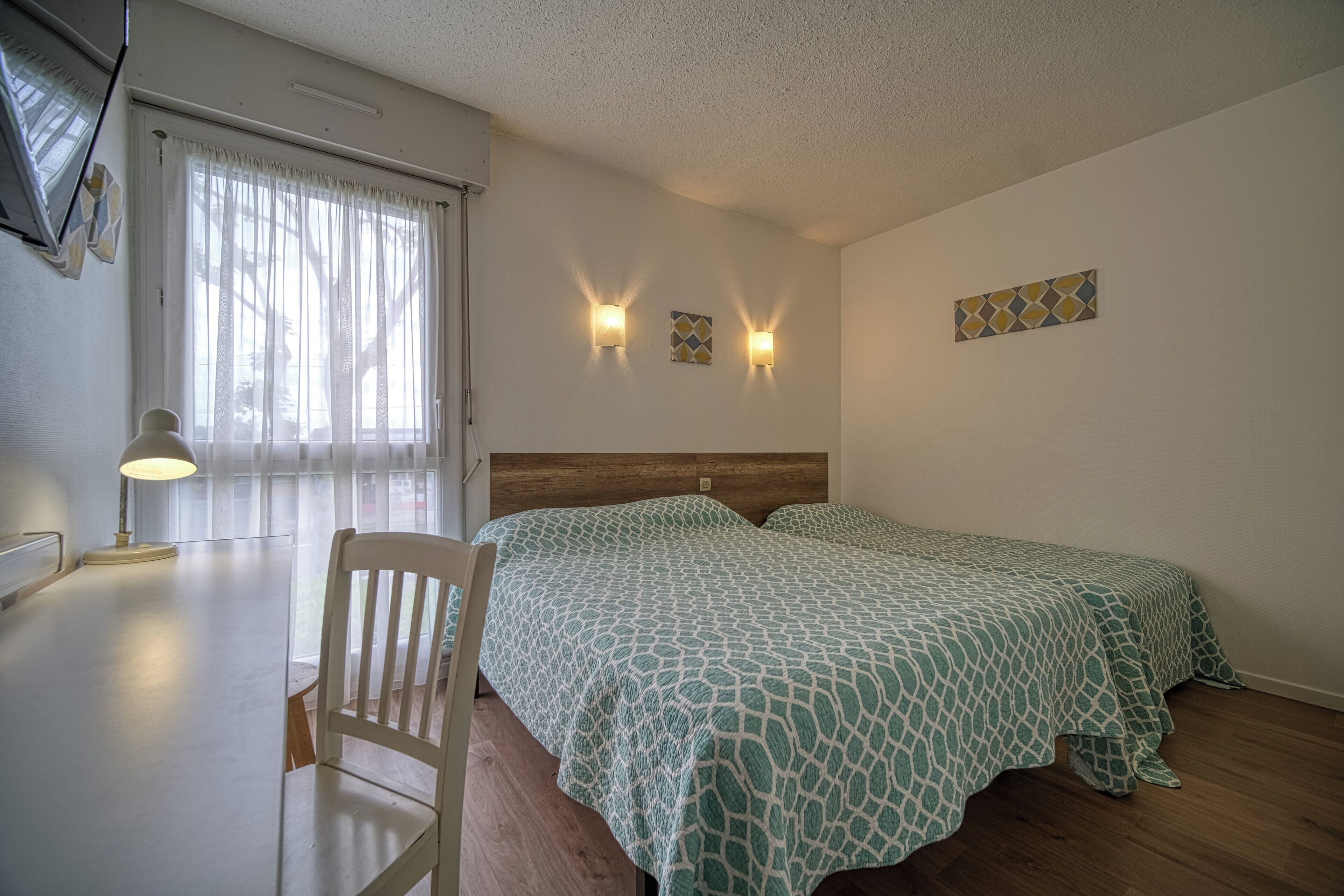 A bedroom with two beds, a desk, and a chair. There are two wall-mounted lights and a window with curtains.