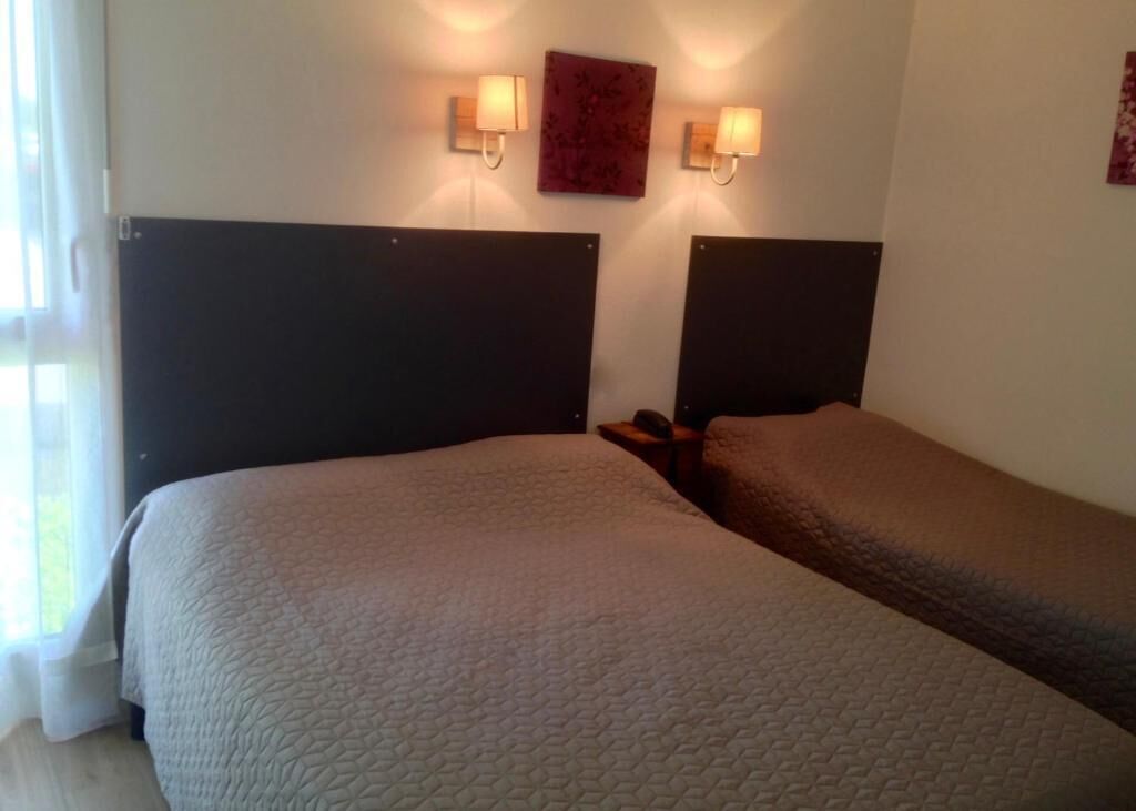 A hotel room with two beds, each with a headboard and a bedside table. There is a wall-mounted light fixture above the beds and a framed picture on the wall.