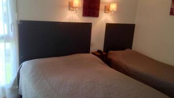 A hotel room with two beds, each with a headboard and a bedside table. There is a wall-mounted light fixture above the beds and a framed picture on the wall.