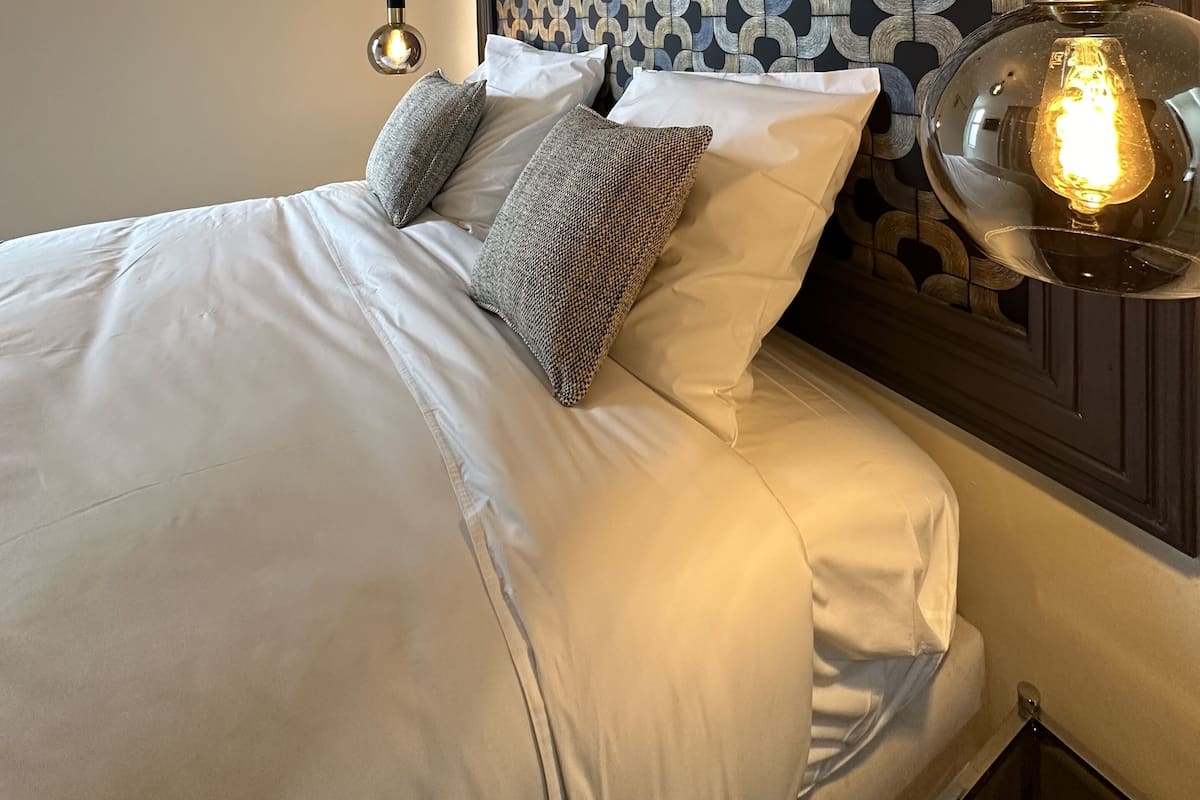 A neatly made bed with white linens and gray pillows, a patterned headboard, and a hanging light fixture.