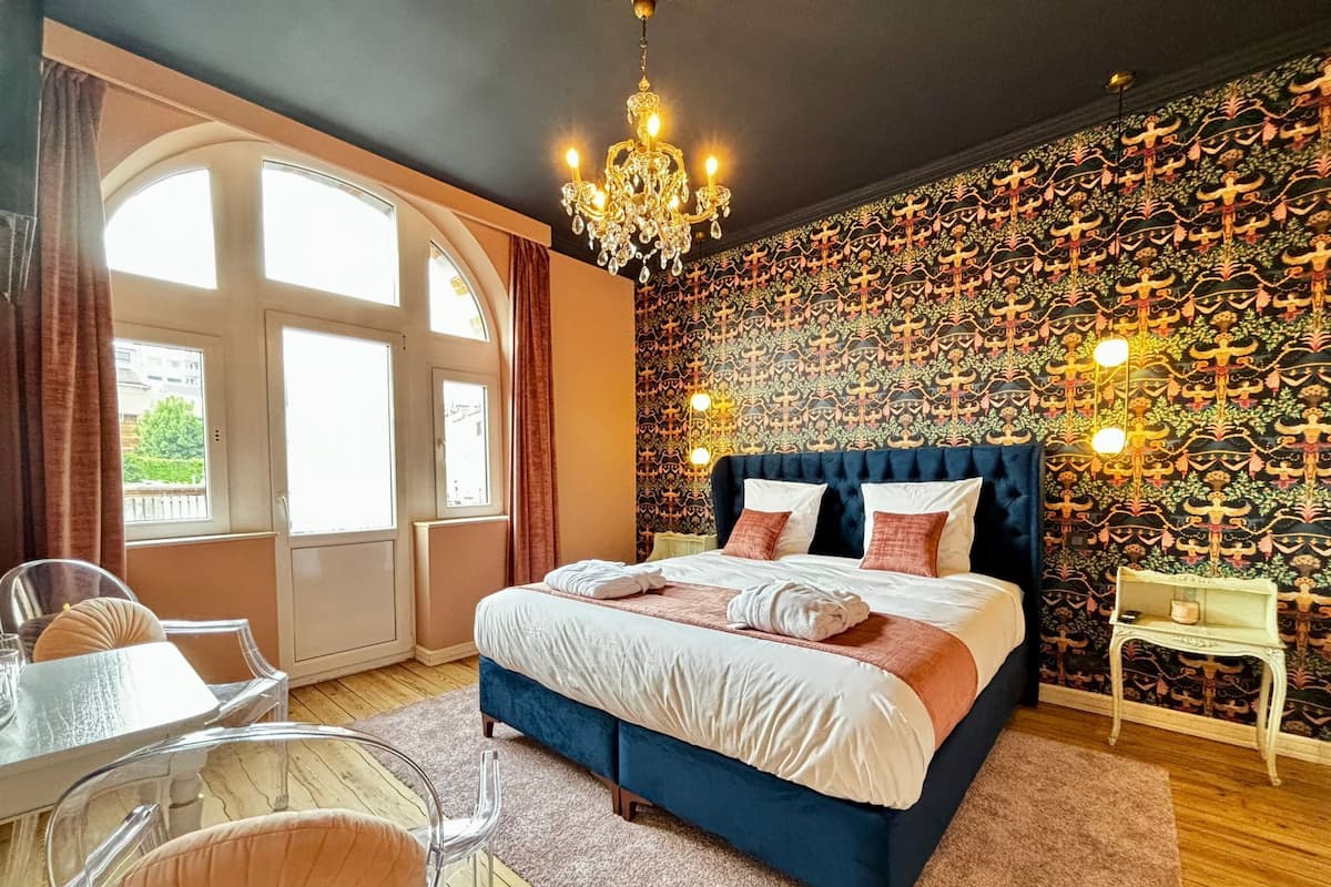 A bedroom with a large bed, a chandelier, a patterned wall, and a view through a window.