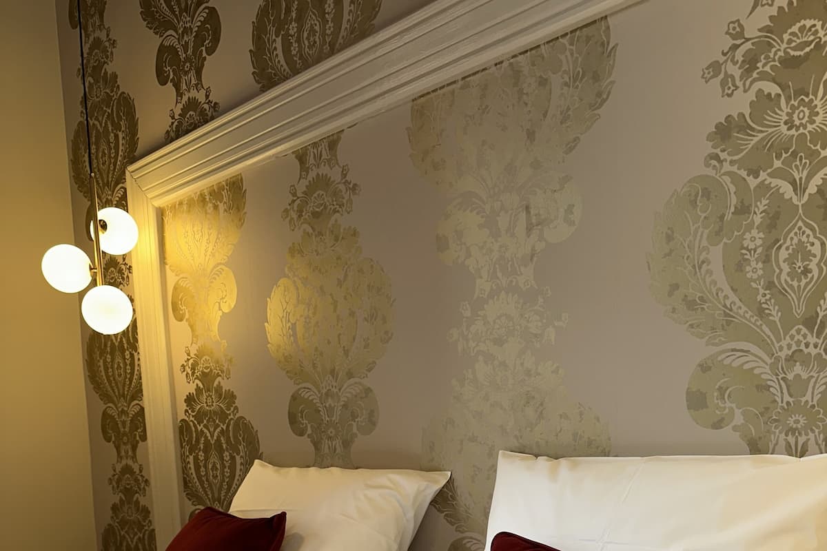 A double bed with white linens and two red pillows, a wall with a floral pattern, and a light fixture with three bulbs.