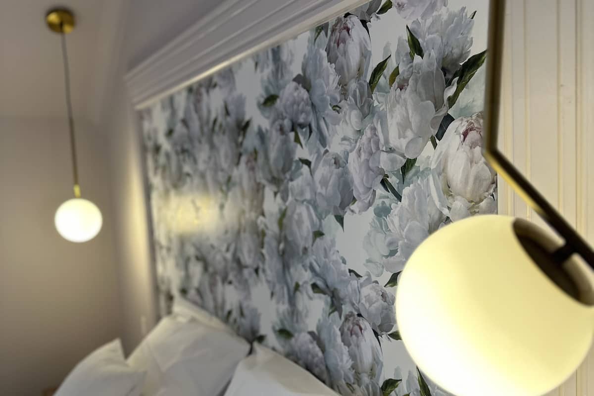 A bedroom with a floral wallpaper, a bed with white linens, and two pendant lights.