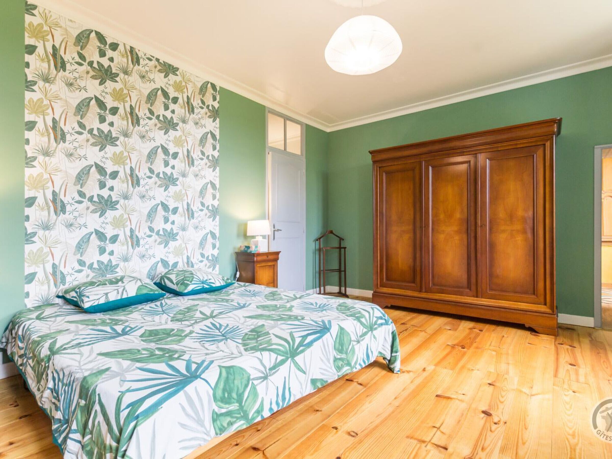 A bedroom with a bed, a wooden wardrobe, a nightstand, and a wall with a floral pattern.