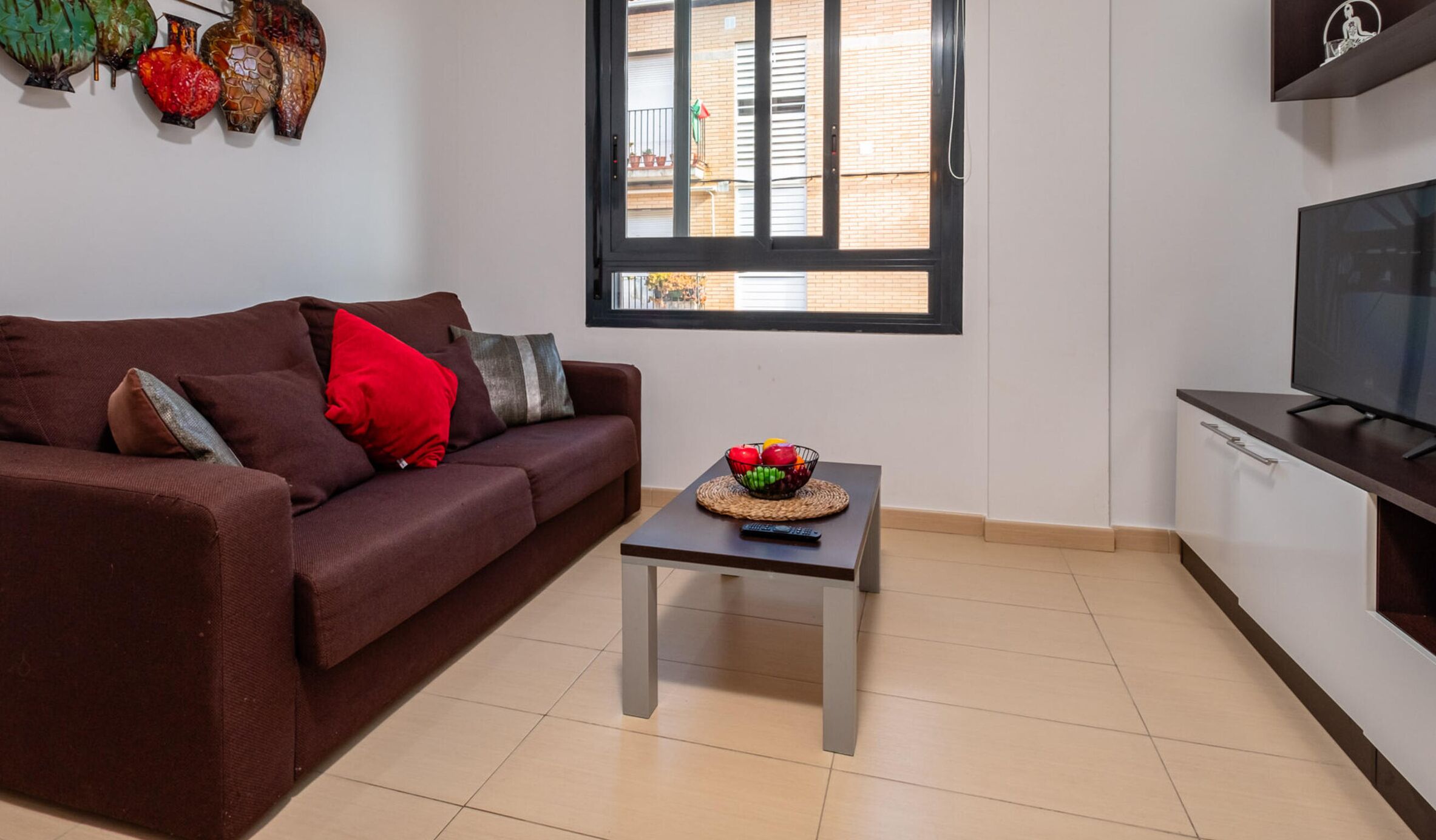 Ein Wohnzimmer mit einem braunen Sofa, einem kleinen Tisch mit Obstschale, einem Fernseher und einem Fenster mit Blick auf ein Gebäude.