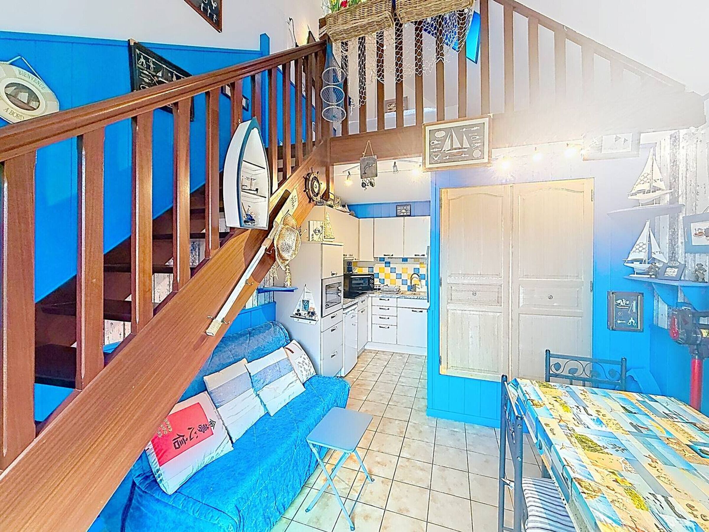 Eine Treppe führt zu einer Küche mit blauer Wand, einem Essbereich mit gemustertem Tischlein und einem blauen Sofa mit Kissen.