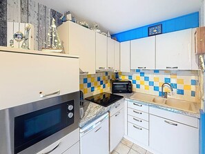 Une cuisine compacte avec des meubles blancs, un micro-ondes noir, un grille-pain et un évier dont le plan de travail est recouvert de carreaux bleus et jaunes.