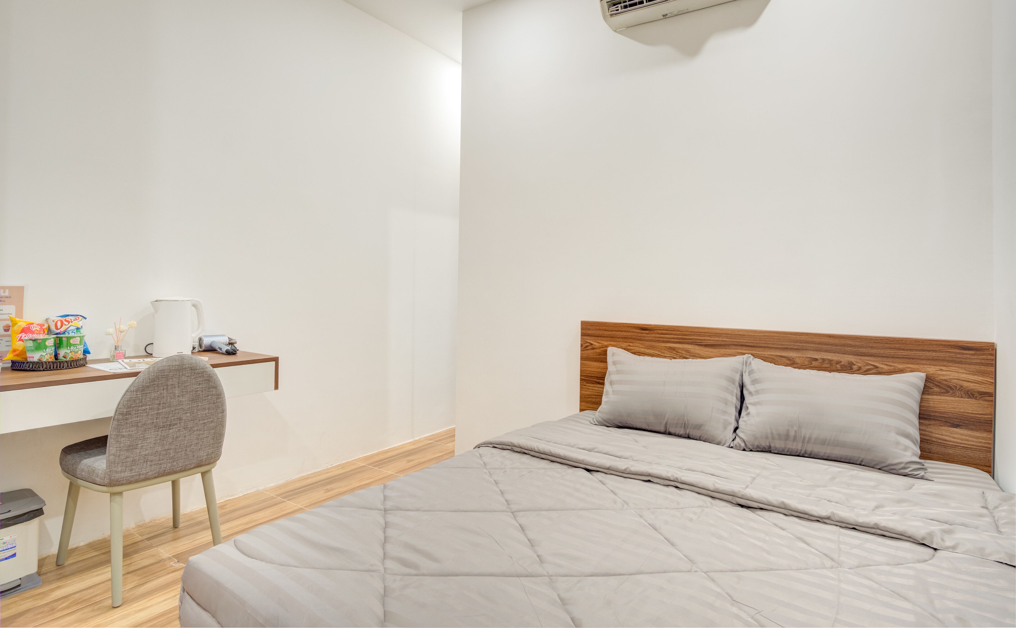 A bedroom with a bed, a wooden headboard, a desk with a chair, a kettle, and some snacks.