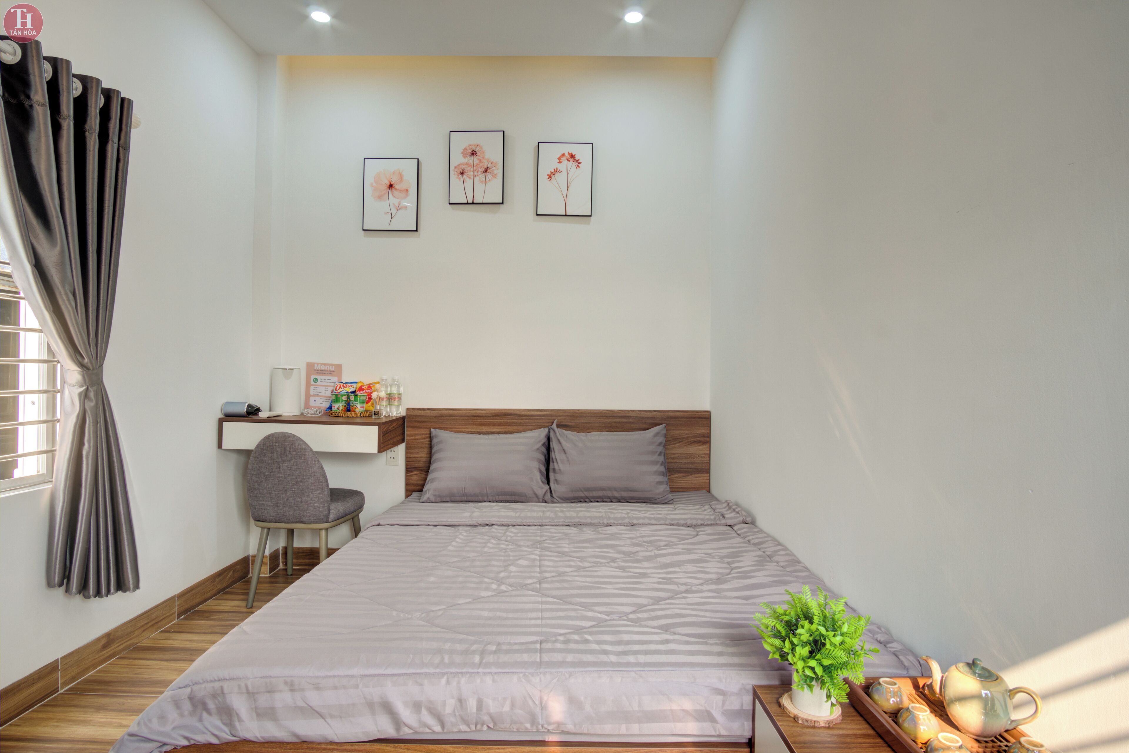Un dormitorio ordenado con cama, escritorio y silla. Hay tres cuadros en la pared, una planta en maceta y una bandeja con teteras y tazas.