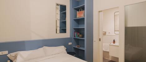 A hotel room with a bed, a shelf with toiletries, a mirror, and a door leading to a bathroom.