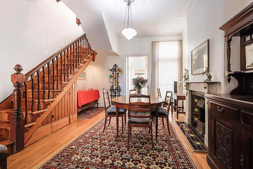 A spacious living area with a wooden staircase, a dining table with chairs, a patterned rug, and a fireplace.