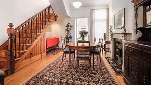A spacious living area with a wooden staircase, a dining table with chairs, a patterned rug, and a fireplace.