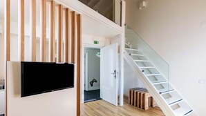 A modern interior with a flat-screen TV mounted on a wall, a wooden slatted partition, a staircase with glass railings, and a door leading to another room. - Porta da Arrábida Suites (Palmela)