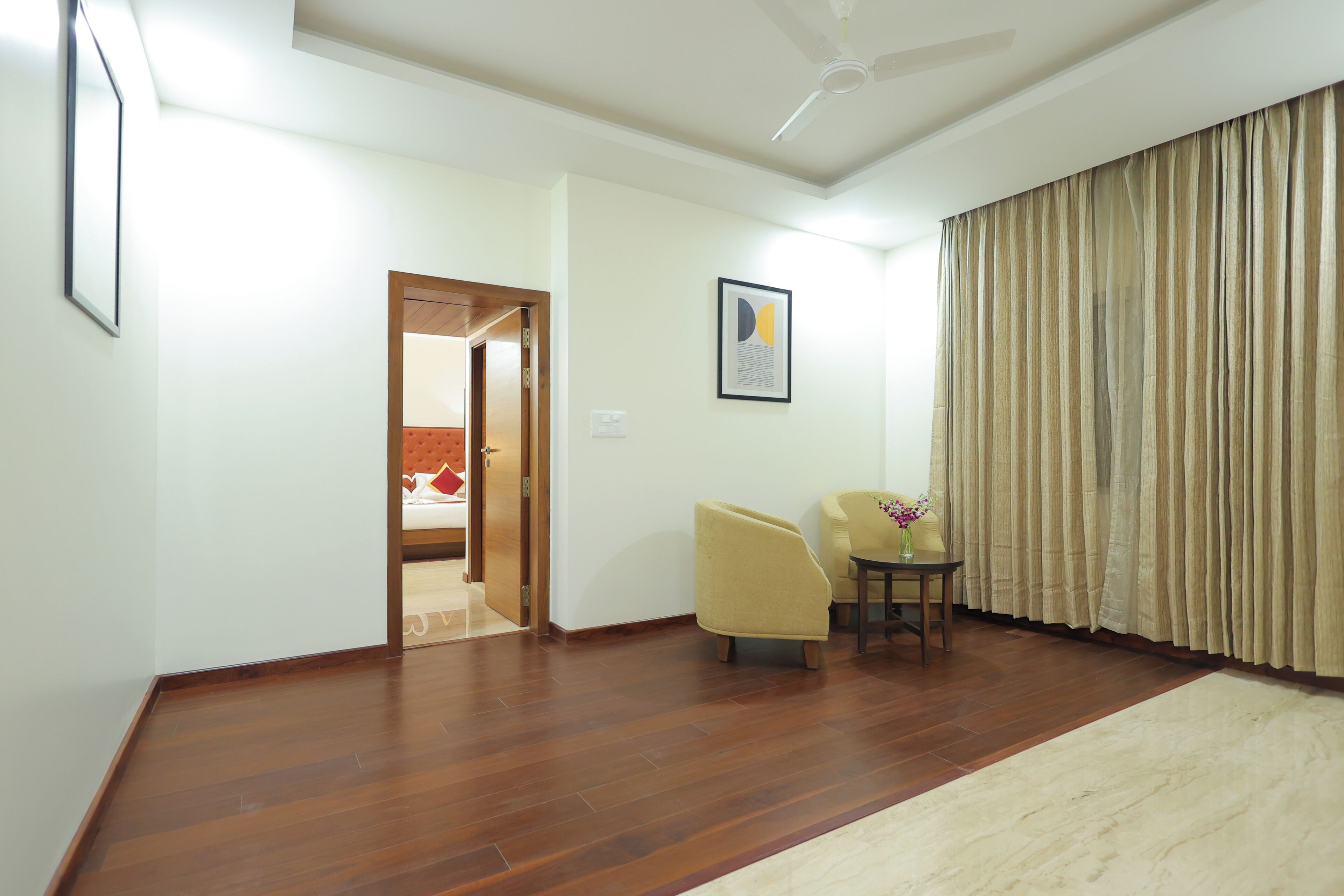 A spacious living room with a wooden floor, a ceiling fan, a door leading to another room, and a beige curtain.