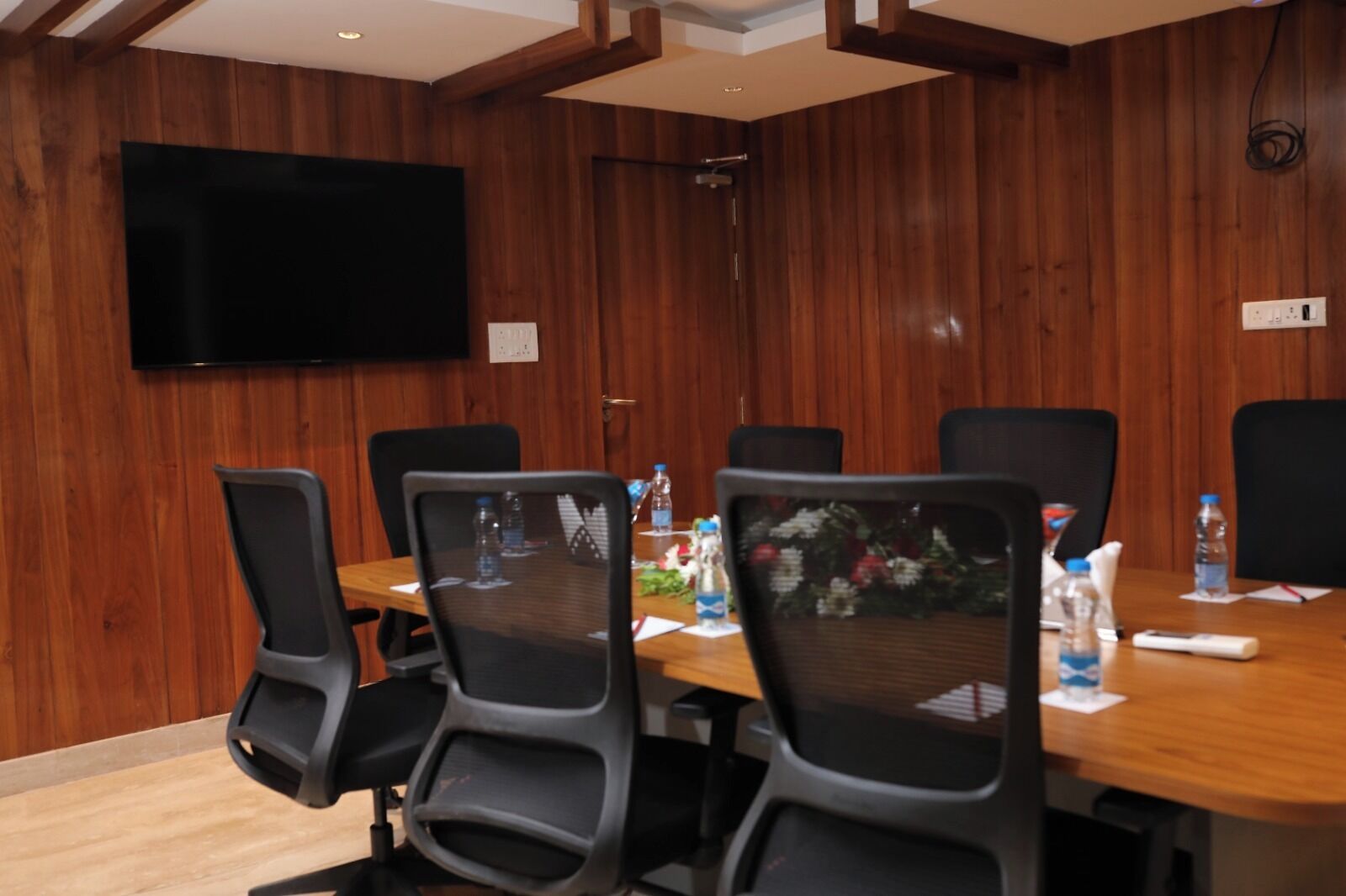 Une salle de conférence aux murs recouverts de panneaux en bois, un téléviseur à écran plat fixé au mur, et une table entourée de chaises et de bouteilles d’eau.