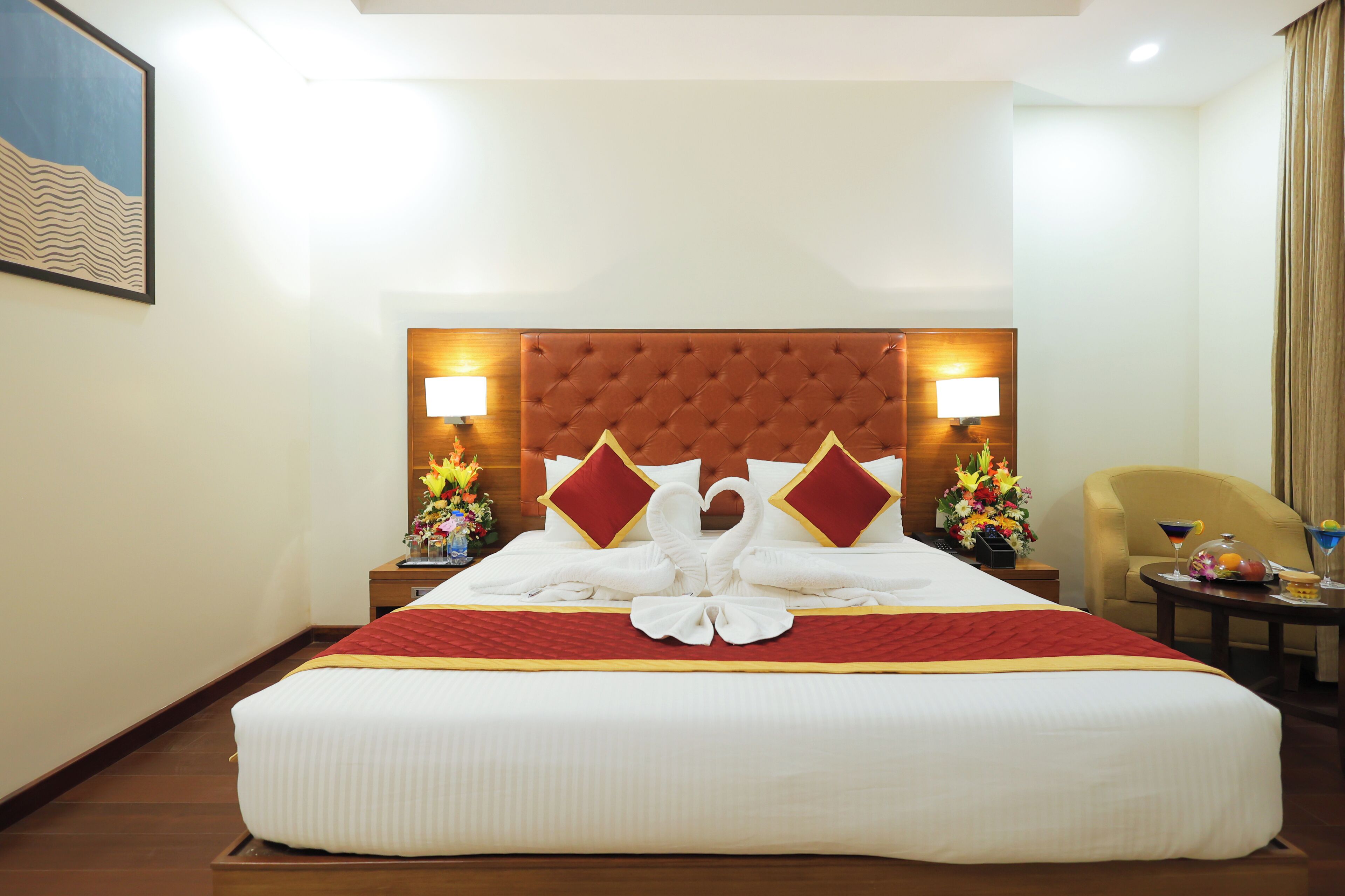 A hotel room with a large bed, a tufted headboard, two bedside lamps, a chair, a small table with a drink and a fruit bowl, and a painting on the wall.