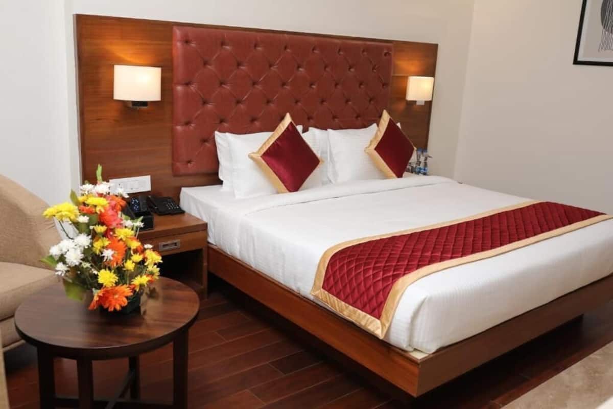 A hotel room with a large bed, a wooden headboard, two bedside lamps, a nightstand with a phone, and a small round table with a vase of flowers.