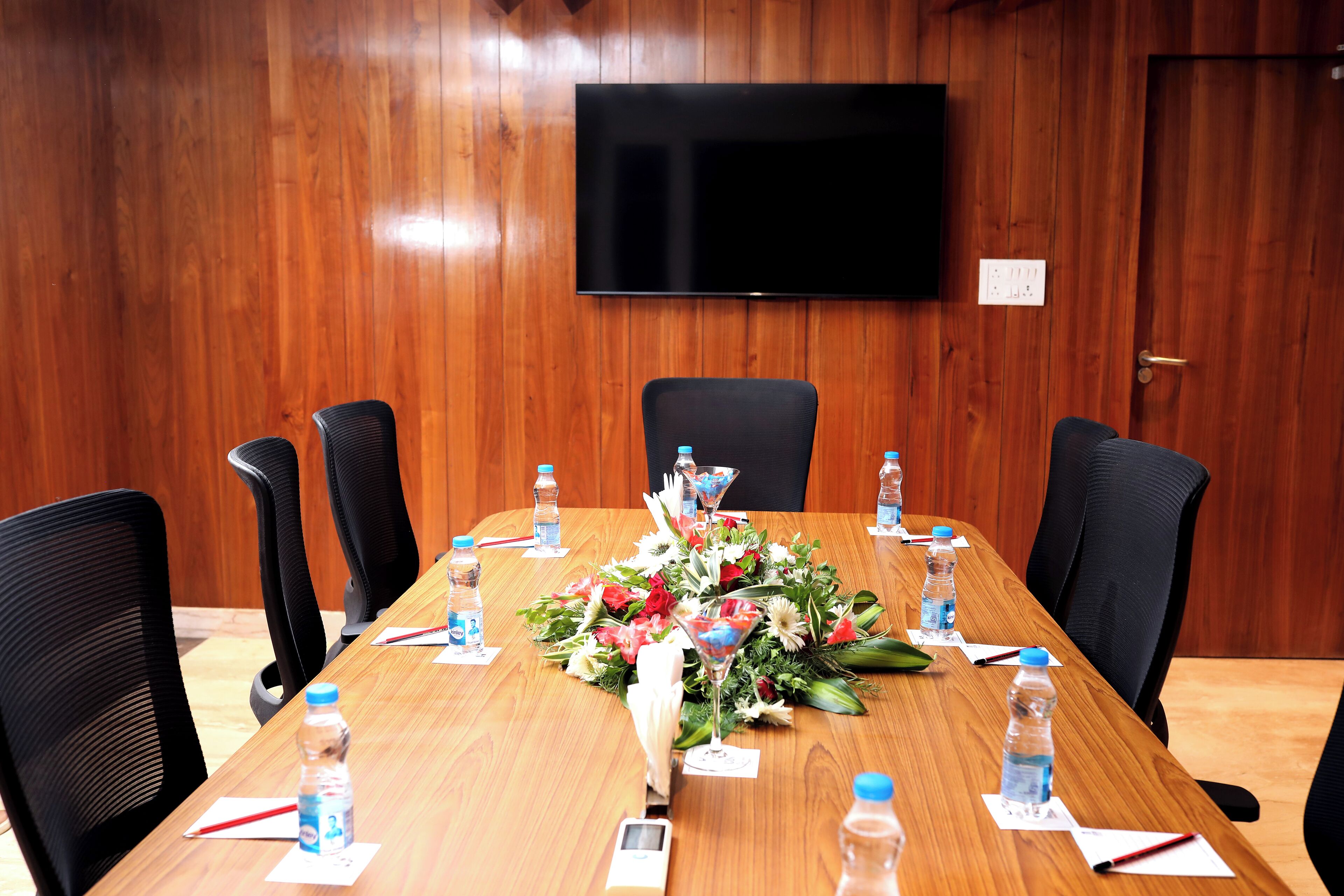 A conference room with a wooden table, black chairs, a floral centerpiece, water bottles, and a TV mounted on the wall.