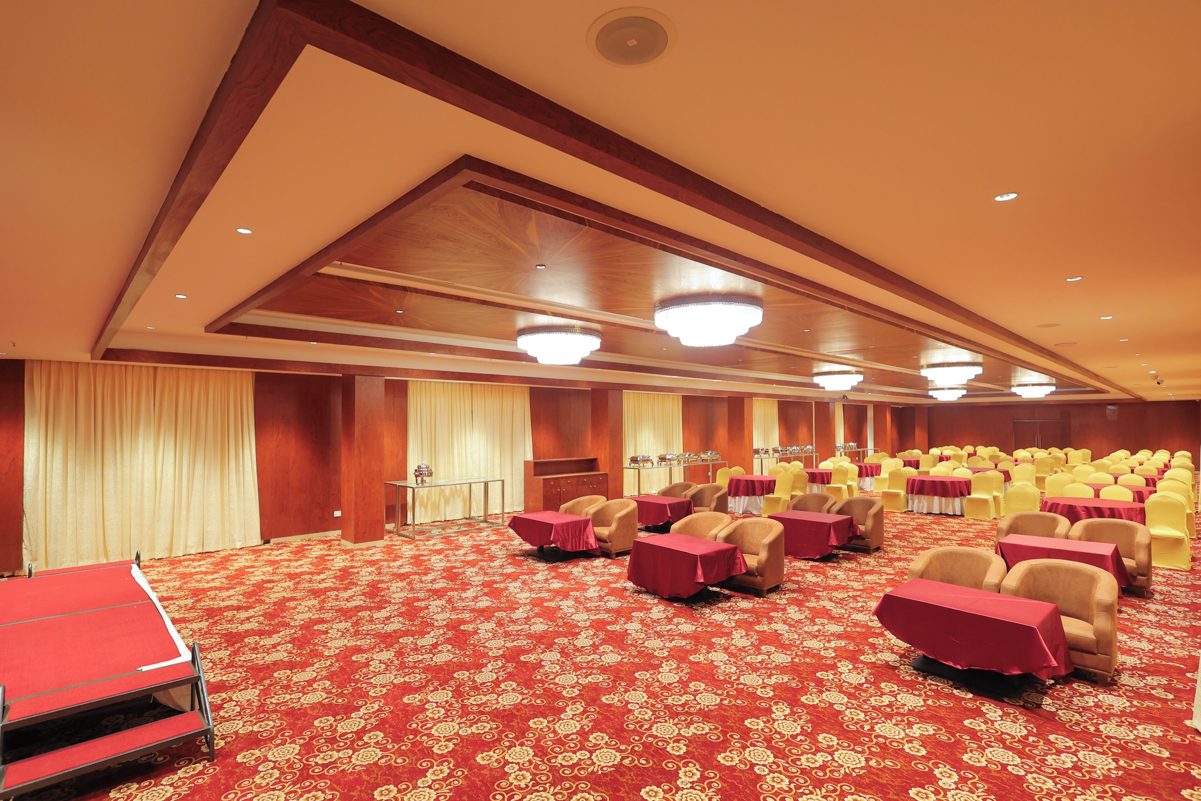 Een ruime congreszaal met rijen gele stoelen en rode tafels, een rood tapijt en houten panelen aan het plafond.