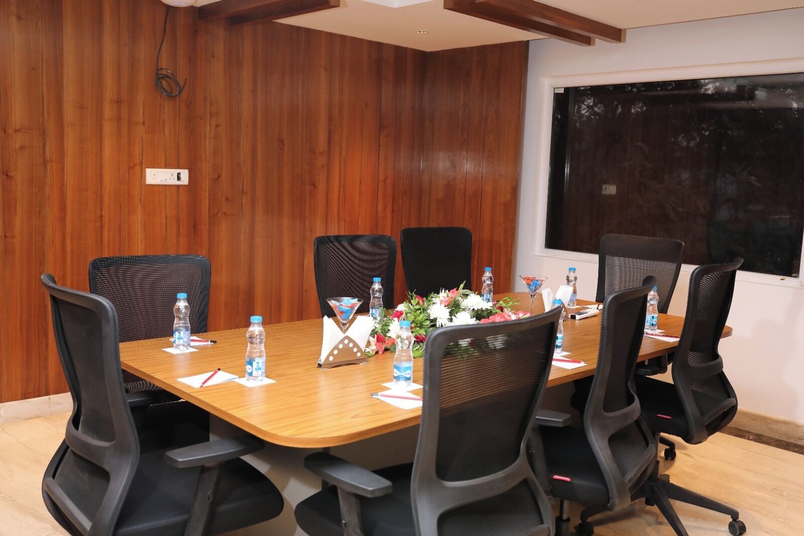 A conference room with a wooden paneled wall, a large table with chairs, water bottles, a flower arrangement, and a projector screen.