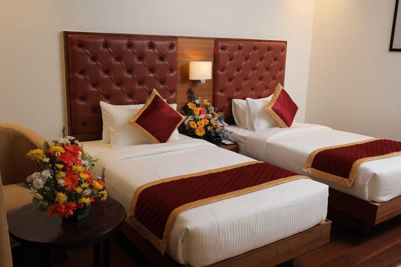 A hotel room with two beds, each with a red headboard, white bedding, and a red throw blanket. There is a small table with a vase of flowers and a lamp on the wall.