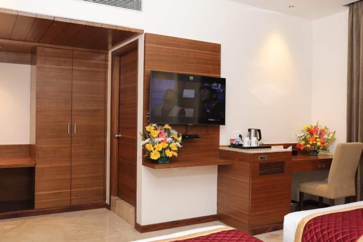 A hotel room with a wooden wardrobe, a TV mounted on the wall, a desk with a chair, and a bed with a red bedspread.