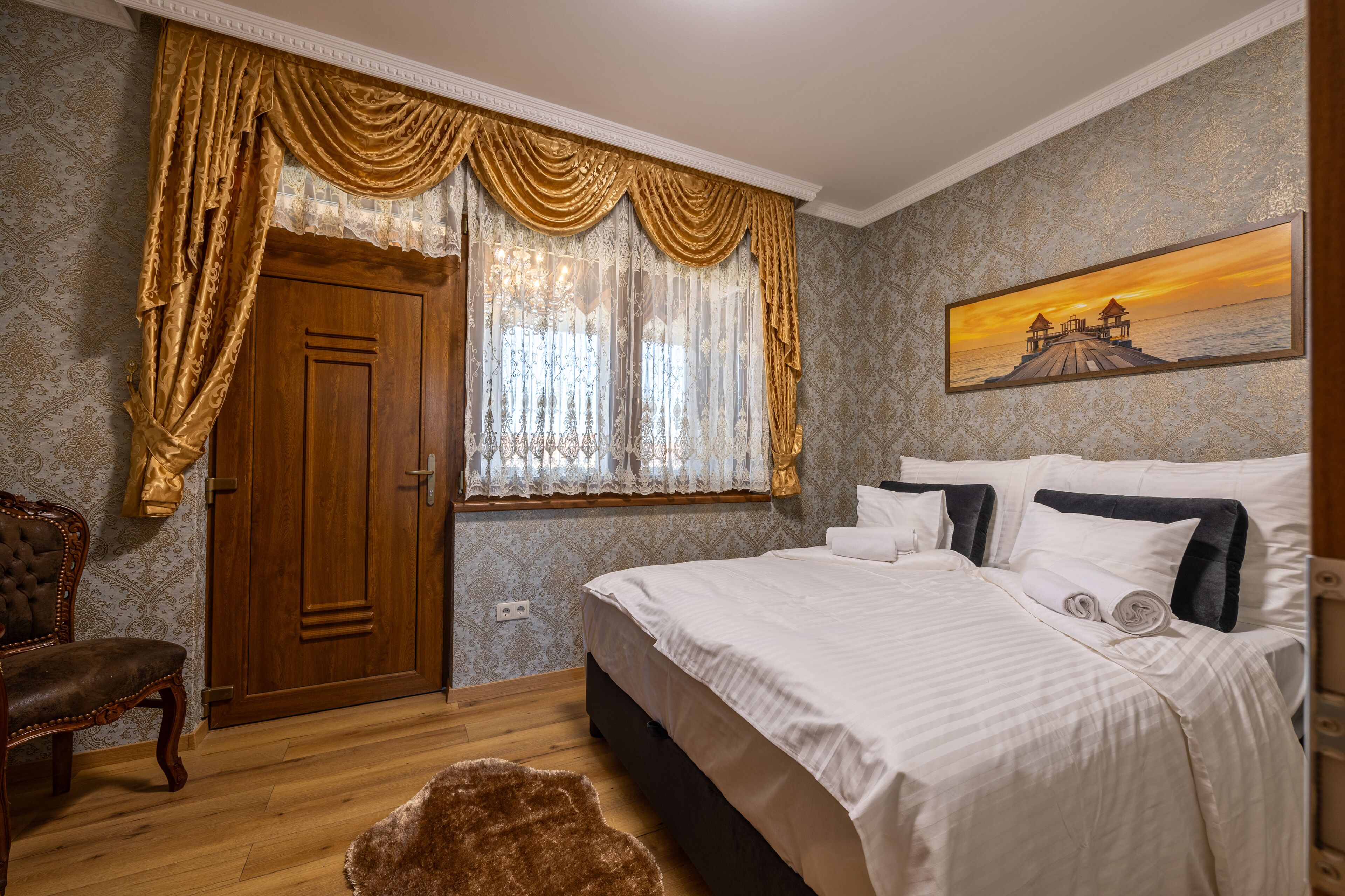 Un dormitorio con puerta de madera, una cama grande con ropa de cama blanca, una silla y un cuadro con atardecer sobre agua.