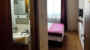 A hotel room with a wooden floor, a bed with a pink bedspread, a television mounted on the wall, and a bathroom with a sink and mirror. - Demirci Otel (Bursa)