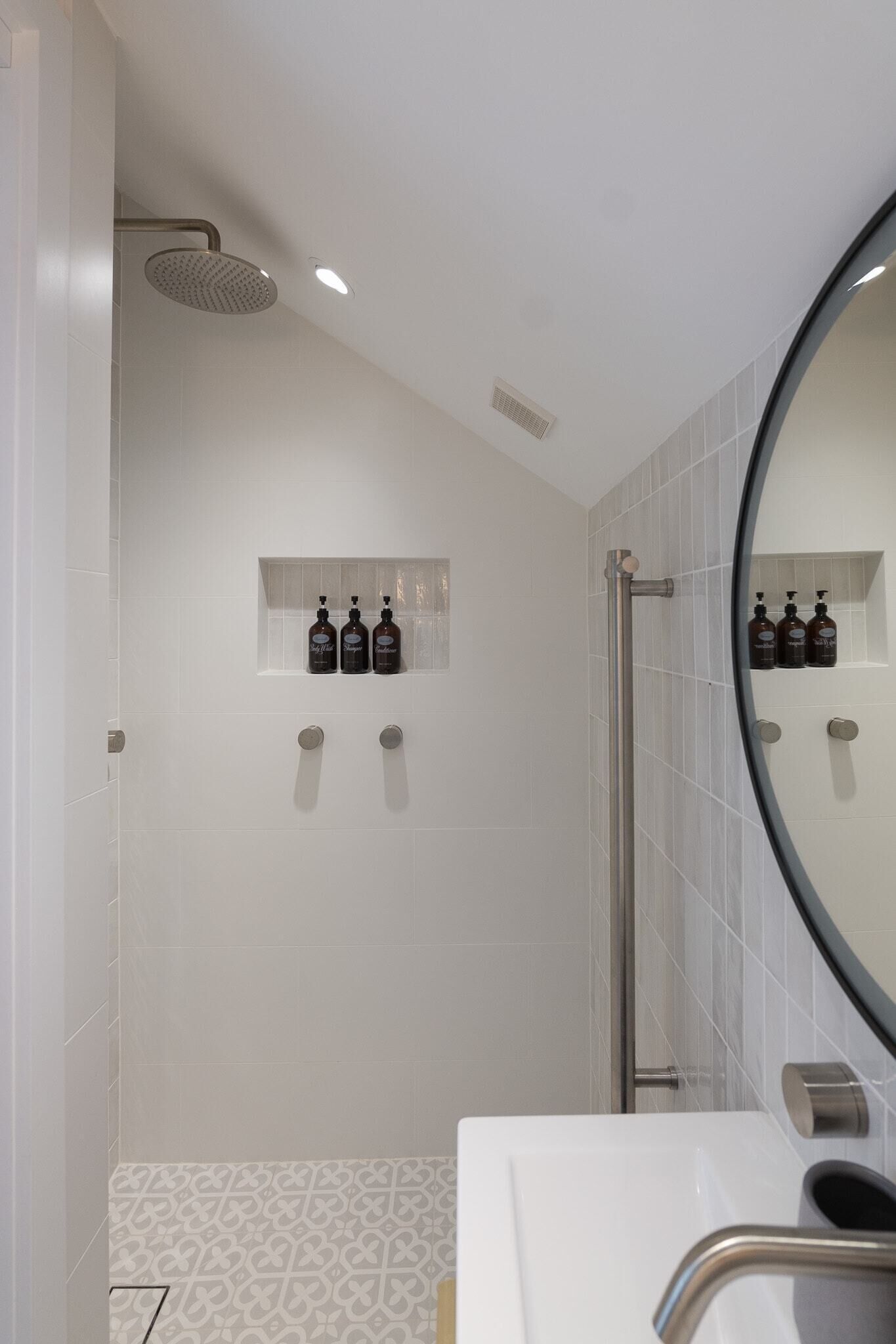 A modern bathroom with a shower, a large mirror, and a shelf with toiletries.