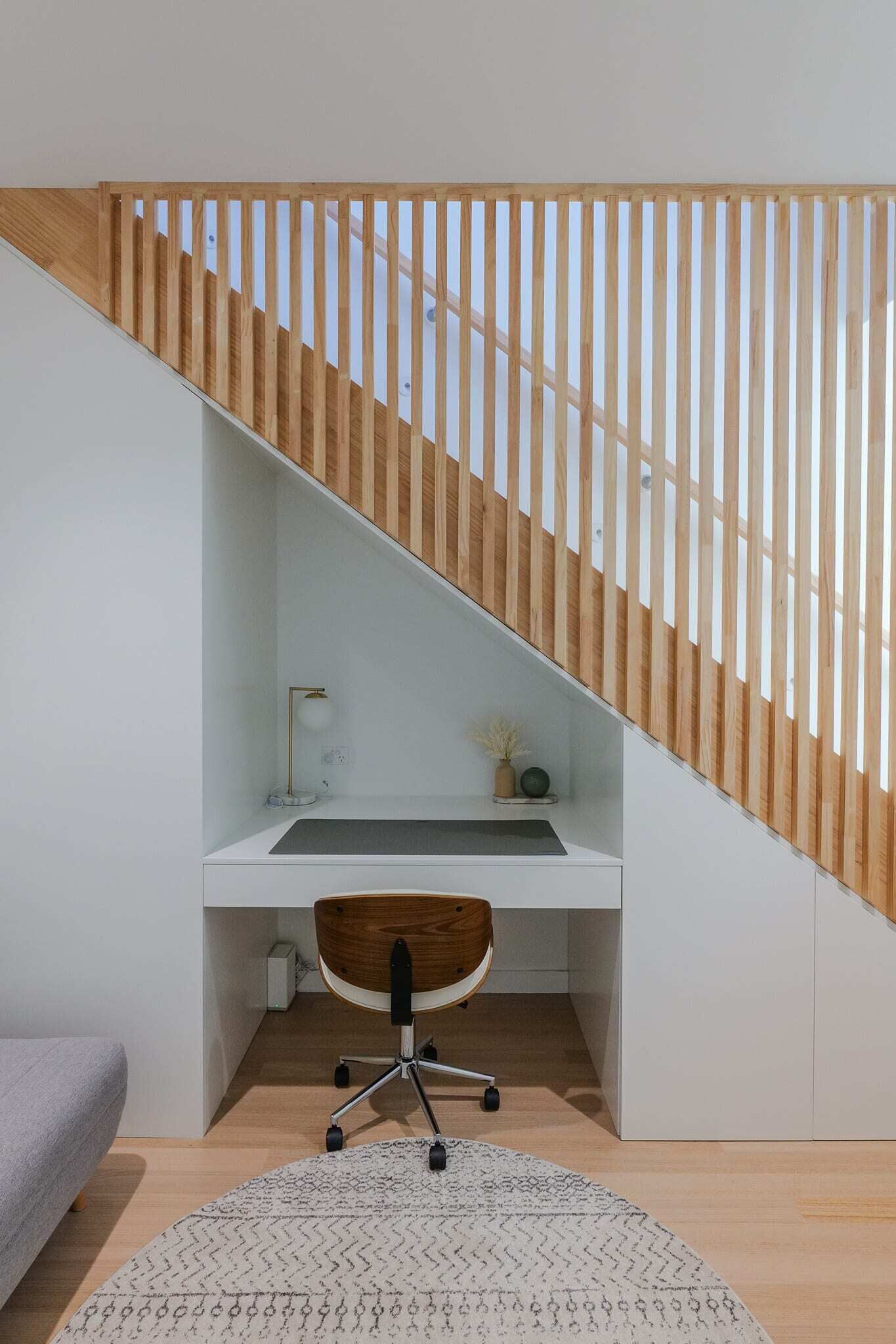 A modern home office with a white desk, a wooden chair, and a staircase with wooden slats.