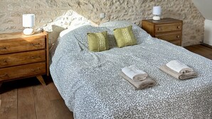A bedroom with a stone wall, a bed with patterned bedding, two pillows, a nightstand with a lamp, and a wooden dresser. - Stylish Country House an hour from Paris (Chevrainvilliers)