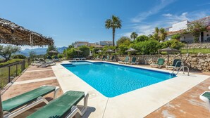 Une piscine rectangulaire à l’eau d’un bleu limpide, entourée de chaises longues et de parasols, avec en toile de fond des montagnes et un ciel dégagé.