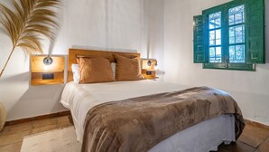 Une chambre avec un lit en bois, deux tables de chevet avec des lampes, une fenêtre avec des volets verts et une grande plante en pot.